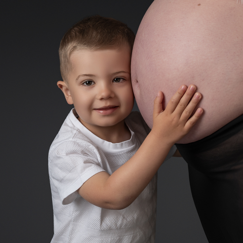 Séance grossesse en famille – Grand frère et ventre rond Petit garçon posant sa joue contre le ventre de sa maman enceinte, séance photo grossesse en famille en studio