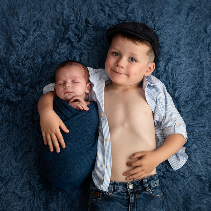 Grand frère allongé sur une fourrure bleue avec son petit frère nouveau-né, séance photo naissance en Vendée