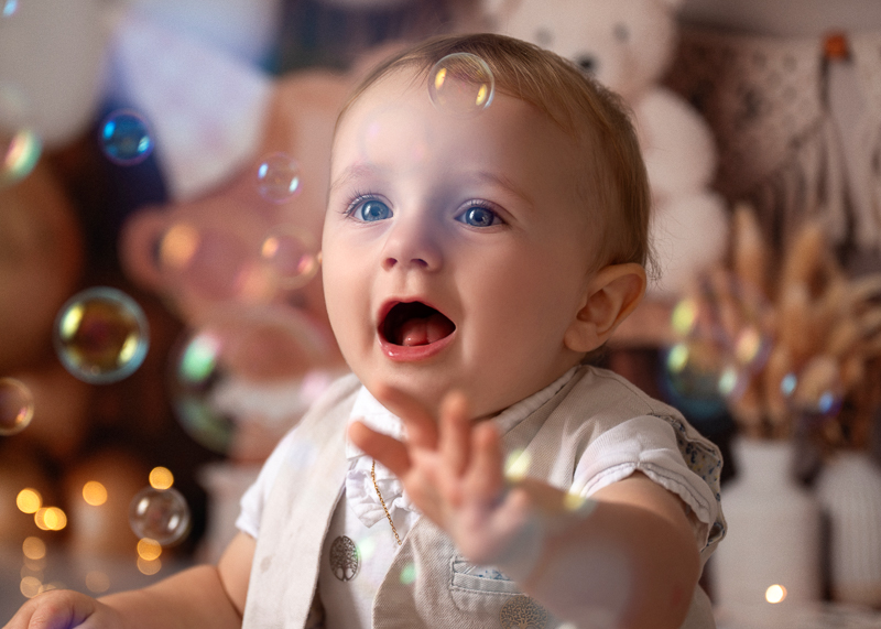 Bébé 12 mois émerveillé par les bulles de savon – La Roche Bébé d'un an bouche ouverte d'émerveillement, tendant la main vers des bulles de savon, dans un décor de studio aux tons clairs avec guirlande lumineuse, séance photo anniversaire bébé près de La Roche-sur-Yon en Vendée