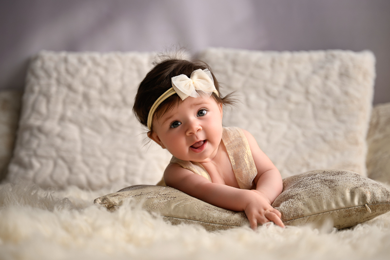 Bébé fille de 4 mois en appui sur les bras, accoudée à un coussin doré, coiffée d'un headband à nœud écru sur un tapis de fourrure blanche, séance photo bébé éveil en studio près de La Chapelle-Hermier en Vendée