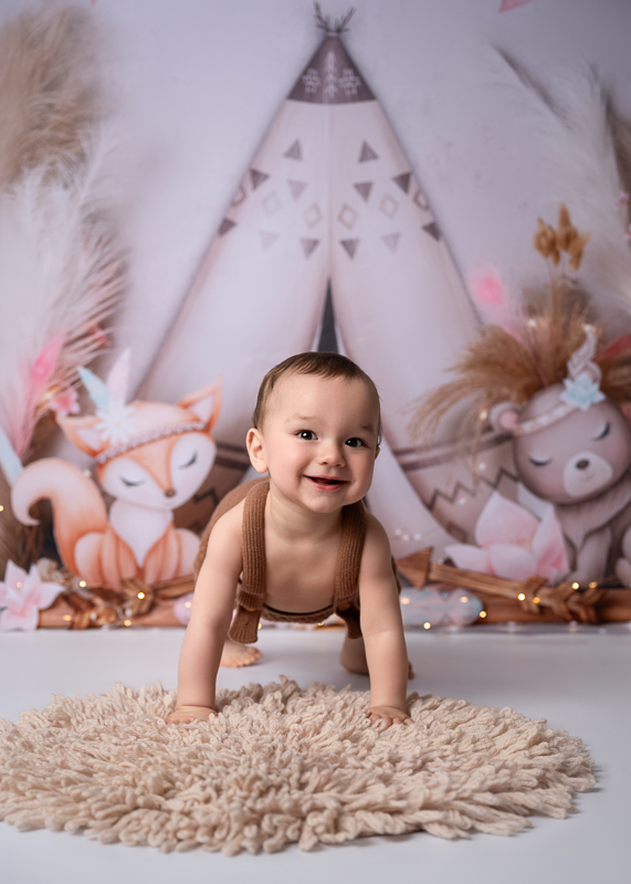 Séance bébé grandit à 9 mois en studio – Le Girouard, Vend Bébé de 9 mois souriant à quatre pattes sur un tapis moelleux, devant un décor de tipi aux illustrations de renard et d'ourson, séance photo bébé grandit en studio près du Girouard en Vendée