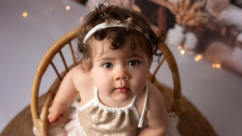 Bébé 9 mois en tenue pailletée, séance bébé grandit – La Bébé fille de 9 mois assise dans un petit fauteuil en rotin, vêtue d'une tenue dorée à paillettes et d'un headband, regard levé vers l'objectif dans un décor lumineux, séance photo bébé grandit en studio près de La Chapelle-Hermier en Vendée