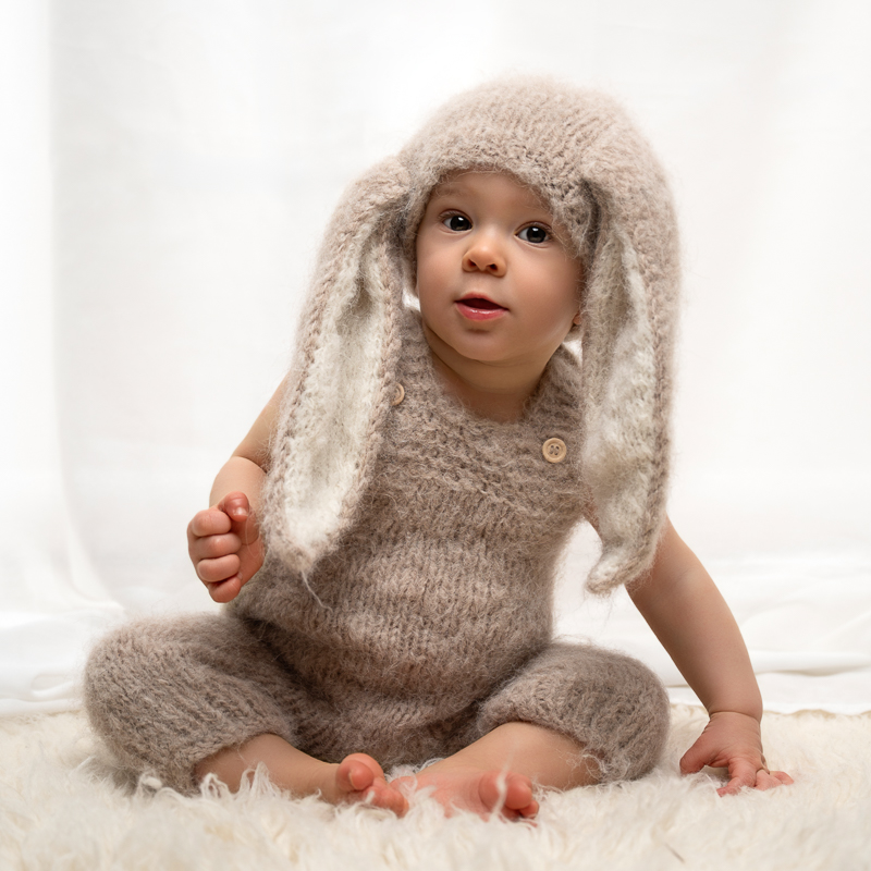 Bébé 9 mois en tenue de lapin, séance bébé grandit – Aize Bébé de 9 mois assis sur une fourrure blanche, vêtu d'un ensemble tricoté beige avec bonnet à grandes oreilles de lapin, séance photo bébé grandit en studio près d'Aizenay en Vendée