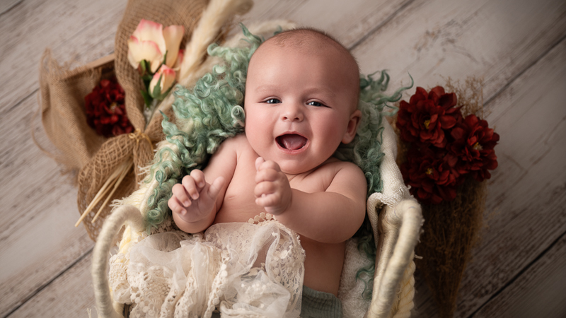 Bébé éveillé riant aux éclats, allongé dans un panier garni de laine bouclée vert d'eau et de dentelle, décor fleuri sur fond de bois clair, séance photo bébé éveil en studio en Vendée