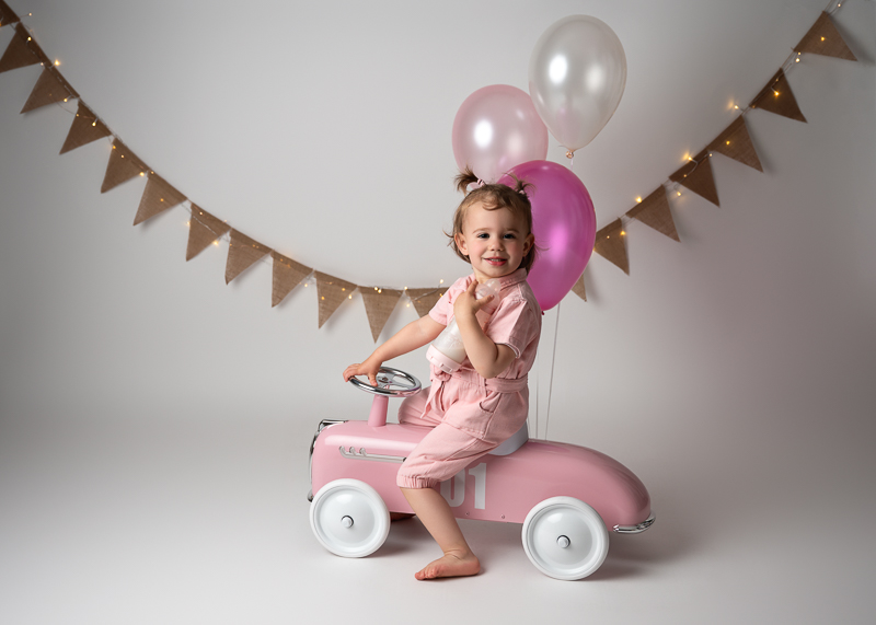 Petite fille de 2 ans souriante assise sur une voiture à pédales rose vintage, tenant son biberon, devant une guirlande de fanions et des ballons roses et blancs, séance photo anniversaire enfant en studio aux Clouzeaux en Vendée