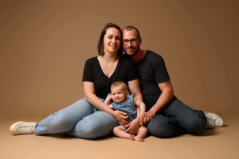 Jeunes parents assis au sol sur fond beige, tenant leur bébé souriant entre eux, tenues assorties jean et t-shirt noir, séance photo famille en studio près de Château-Guibert en Vendée