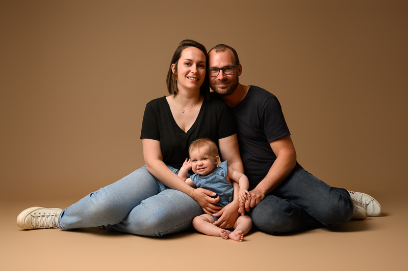 Jeunes parents assis au sol sur fond beige, tenant leur bébé souriant entre eux, tenues assorties jean et t-shirt noir, séance photo famille en studio près de Château-Guibert en Vendée