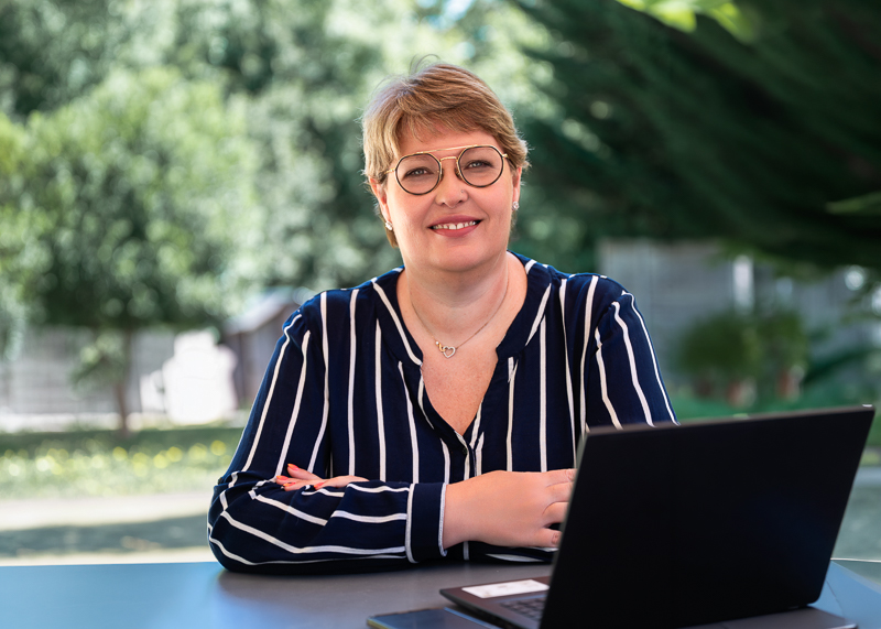 Femme entrepreneure souriante installée à un bureau extérieur devant son ordinateur portable, chemisier rayé bleu marine et verdure en arrière-plan, séance photo portrait professionnel à Aubigny-Les-Clouzeaux en Vendée