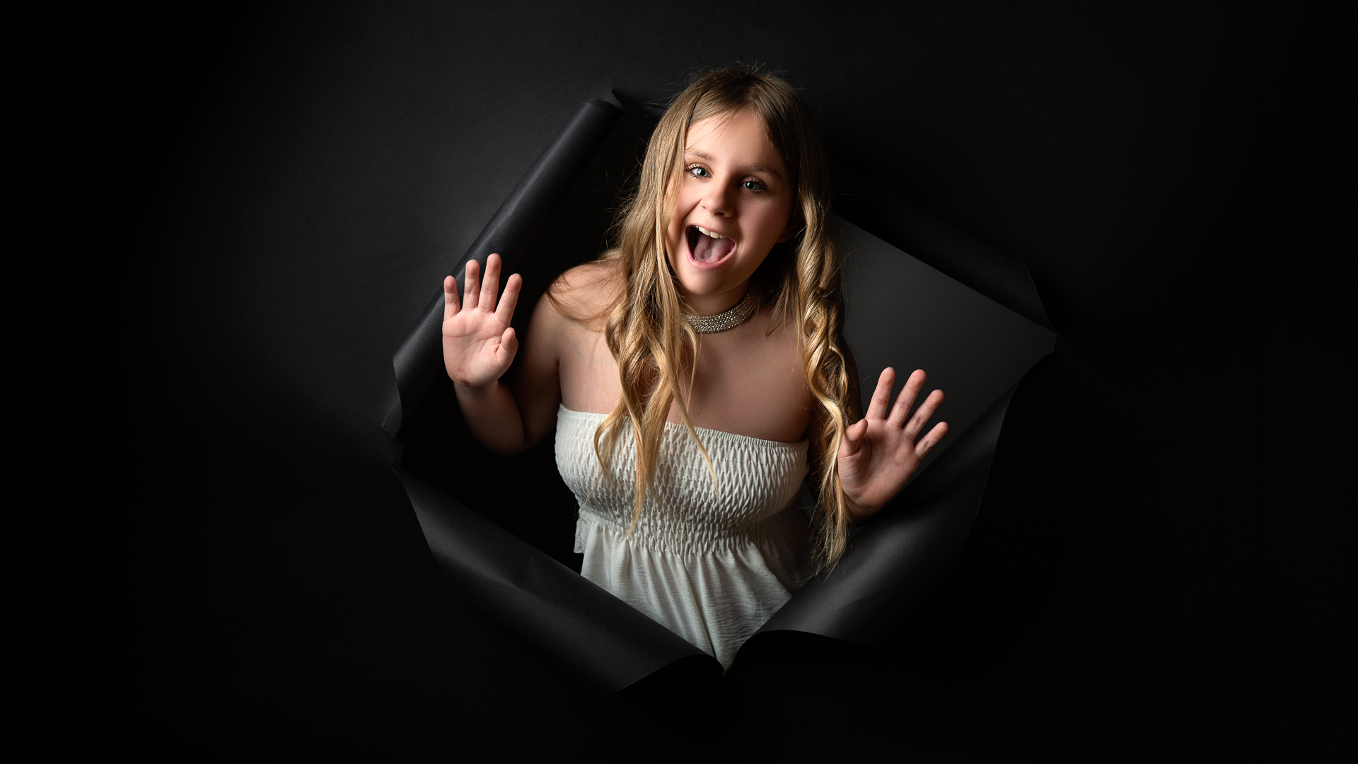 Adolescente souriante surgissant d'un fond noir déchiré, mains levées et cheveux blonds ondulés, portrait créatif réalisé en studio près de La Roche-sur-Yon en Vendée