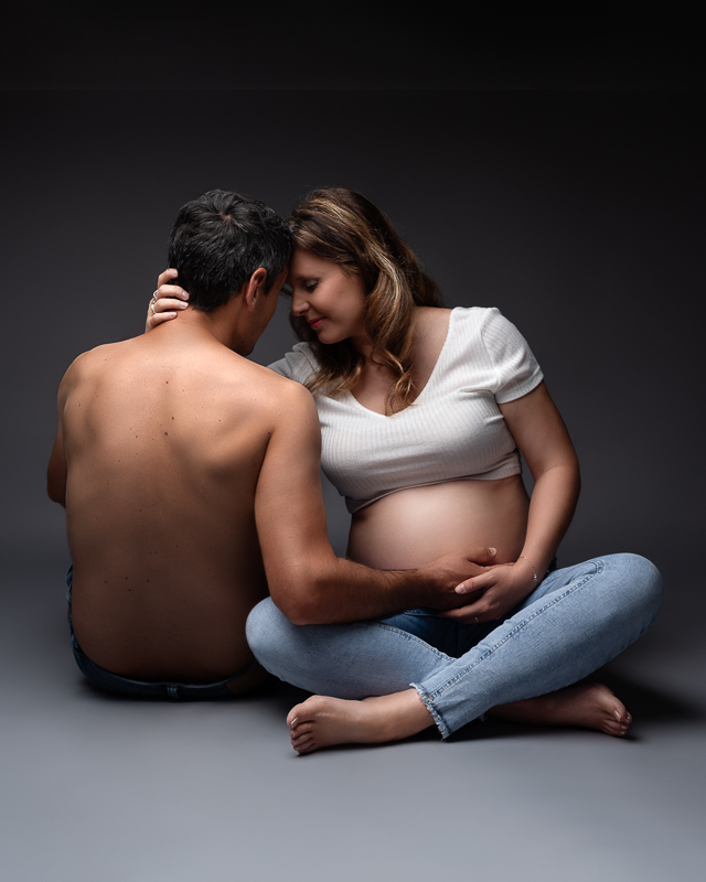 Futurs parents assis au sol front contre front, mains sur le ventre rond, jean et top blanc, photo de grossesse en studio aux Clouzeaux