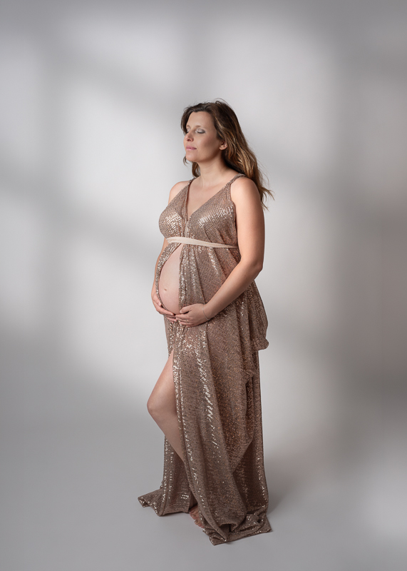 Future maman en robe longue ouverte à sequins dorés, yeux fermés, mains sur le ventre, séance photo grossesse en studio en Vendée
