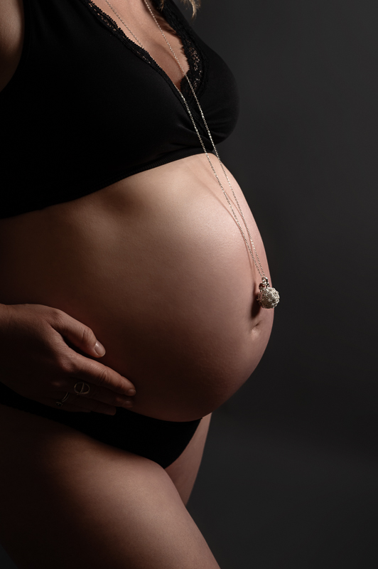 Ventre rond de profil avec un bola de grossesse en pendentif, lingerie noire sur fond sombre, photographe grossesse en Vendée