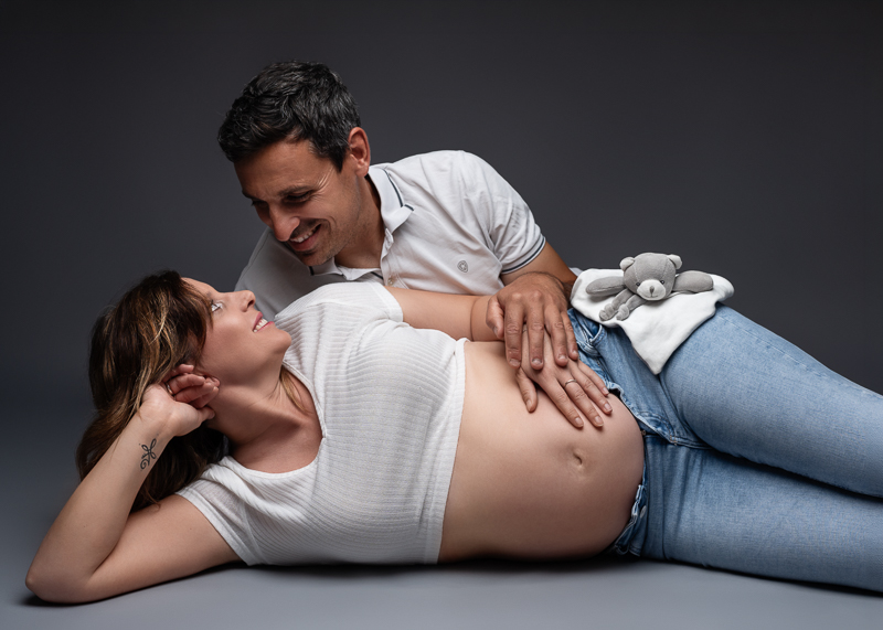 Futur papa souriant, main sur le ventre de la future maman allongée, doudou posé à côté, séance grossesse en couple pour une famille de Sainte-Flaive-des-Loups